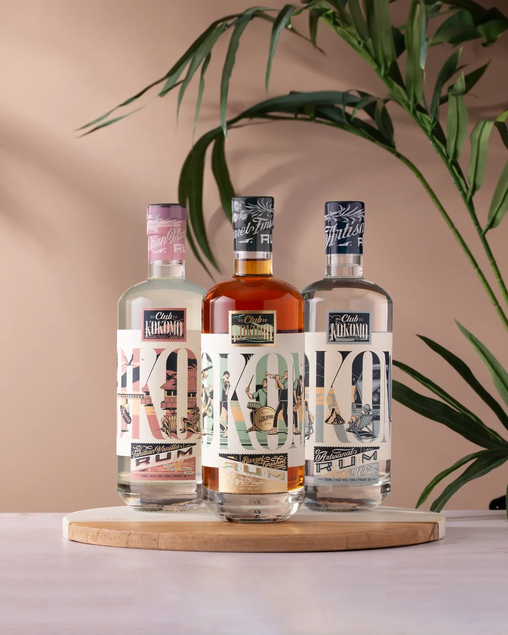 Three Club Kokomo rum bottles with distinct pink, black, and blue closures, arranged beside palm leaves
